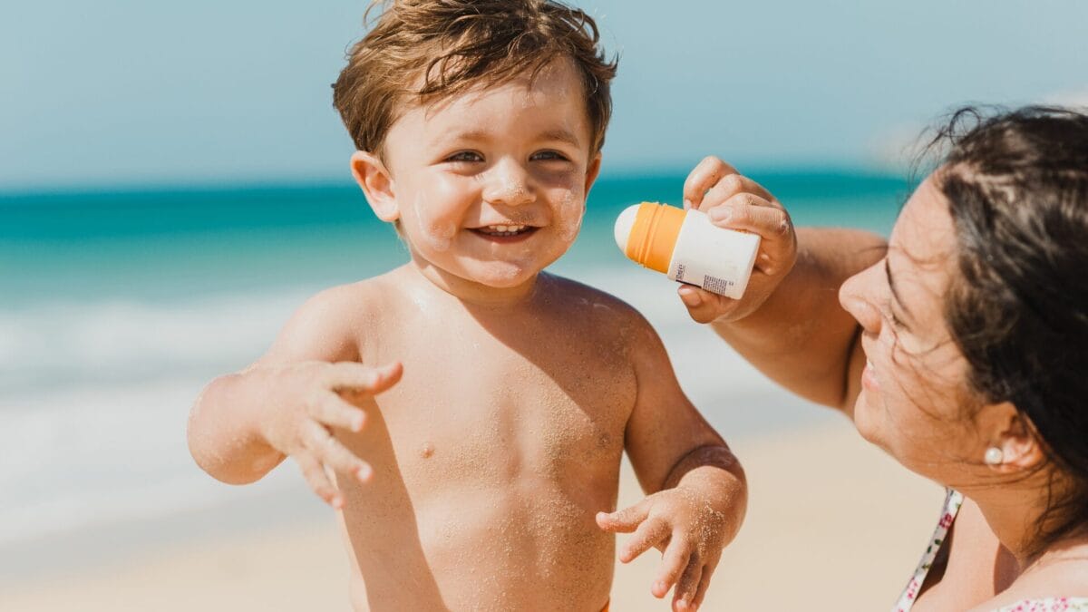 Photo of mom applying sunscreen on toddler by the beach